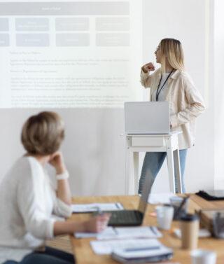 une femme assise regarde vers une femme debout qui présente un powerpoint orienté au mur
